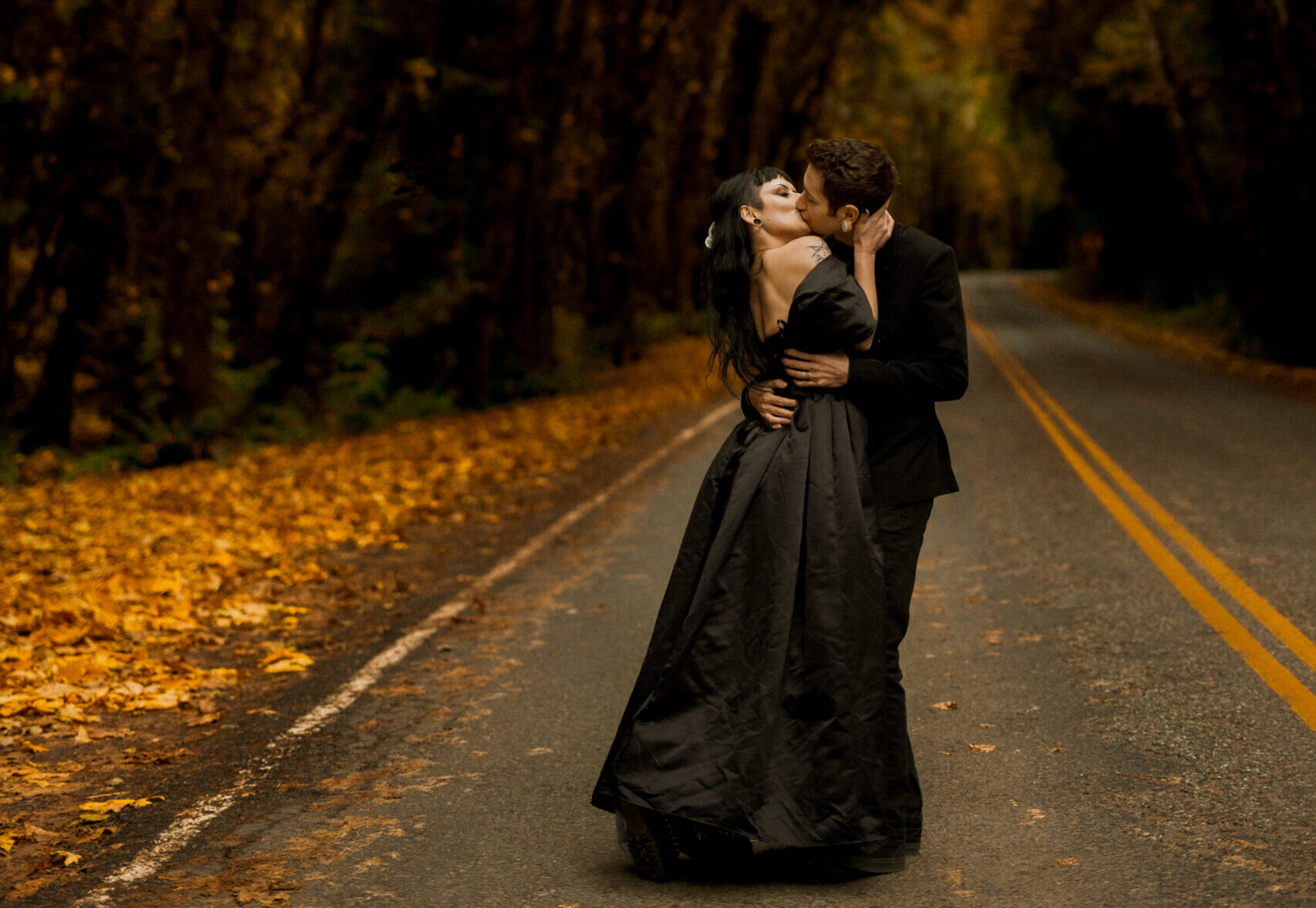 A newly eloped couple wearing all black shares a dip kiss on a fall road in the Hoh Rainforest.