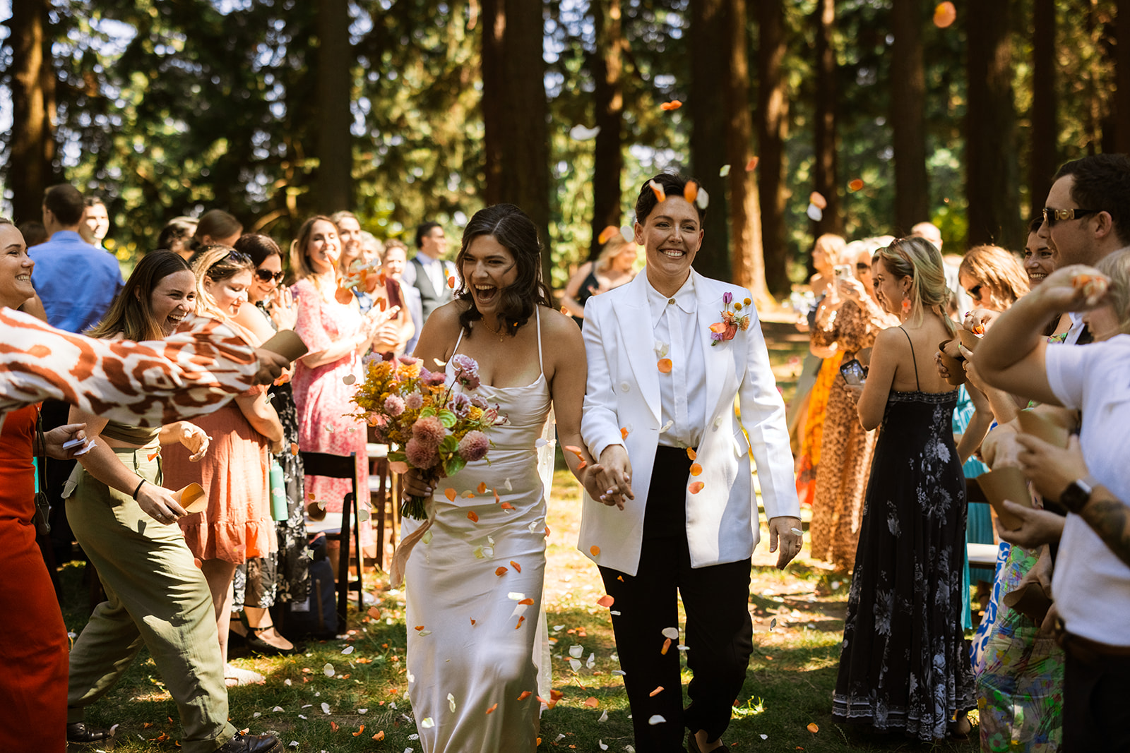 A queer couple walks hand in hand receding down their wedding aisle as their guests throw confetti, cheer, and clap, outside in a Portland park.